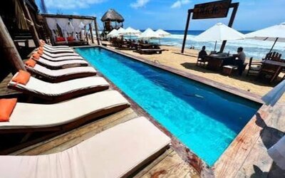 Beachfront swimming pool lined with cushioned wooden sun loungers, with white umbrellas, beach seating and ocean views under a bright blue sky.