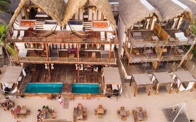 Aerial view of Hotel Nude’s beachfront buildings with thatched roofs, balconies, two rectangular pools, cabanas and dining tables on the sand.