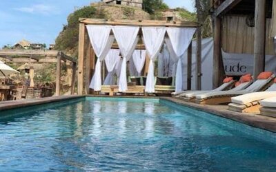 Rectangular outdoor swimming pool with cushioned loungers and a curtained wooden cabana, set beside the hotel near a rocky coastal hillside.