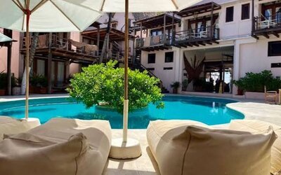 Shaded view across a turquoise courtyard pool from two cushioned lounge chairs, with large white umbrellas and hotel balconies in the background.