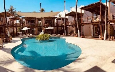 Curved turquoise outdoor swimming pool in a sunny courtyard surrounded by rustic wooden hotel buildings, palm trees and shaded lounge areas.