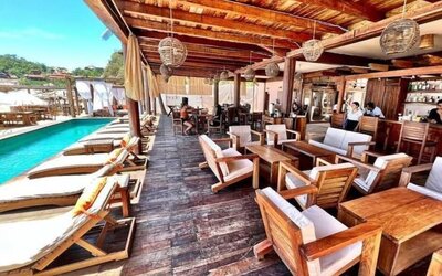 Open-air wooden terrace beside a rectangular swimming pool, featuring cushioned loungers, dining tables, armchairs, pendant lights and a bar.