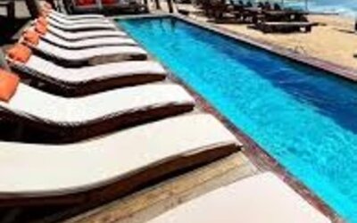 Long beachfront swimming pool lined with cushioned wooden loungers, white umbrellas and seating overlooking the blue ocean.