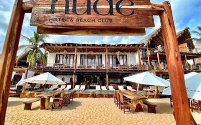 Wooden Hotel Nude Hotel & Beach Club entrance sign framing the beachfront property, with umbrellas, tables and loungers on the sand.