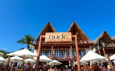 Beachfront view of Hotel Nude with a large wooden entrance sign, thatched-roof architecture, white umbrellas and outdoor seating beneath a bright blue sky.
