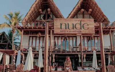 Front exterior of Hotel Nude Hotel & Beach Club with rustic wooden balconies, steep thatched roofs, beach loungers and umbrellas on the sand.
