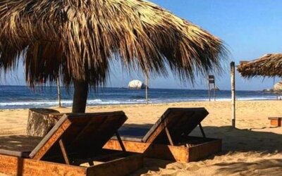 Two wooden sun loungers rest beneath a large thatched palapa on a sandy beach, overlooking the blue ocean and breaking waves under a clear sky.