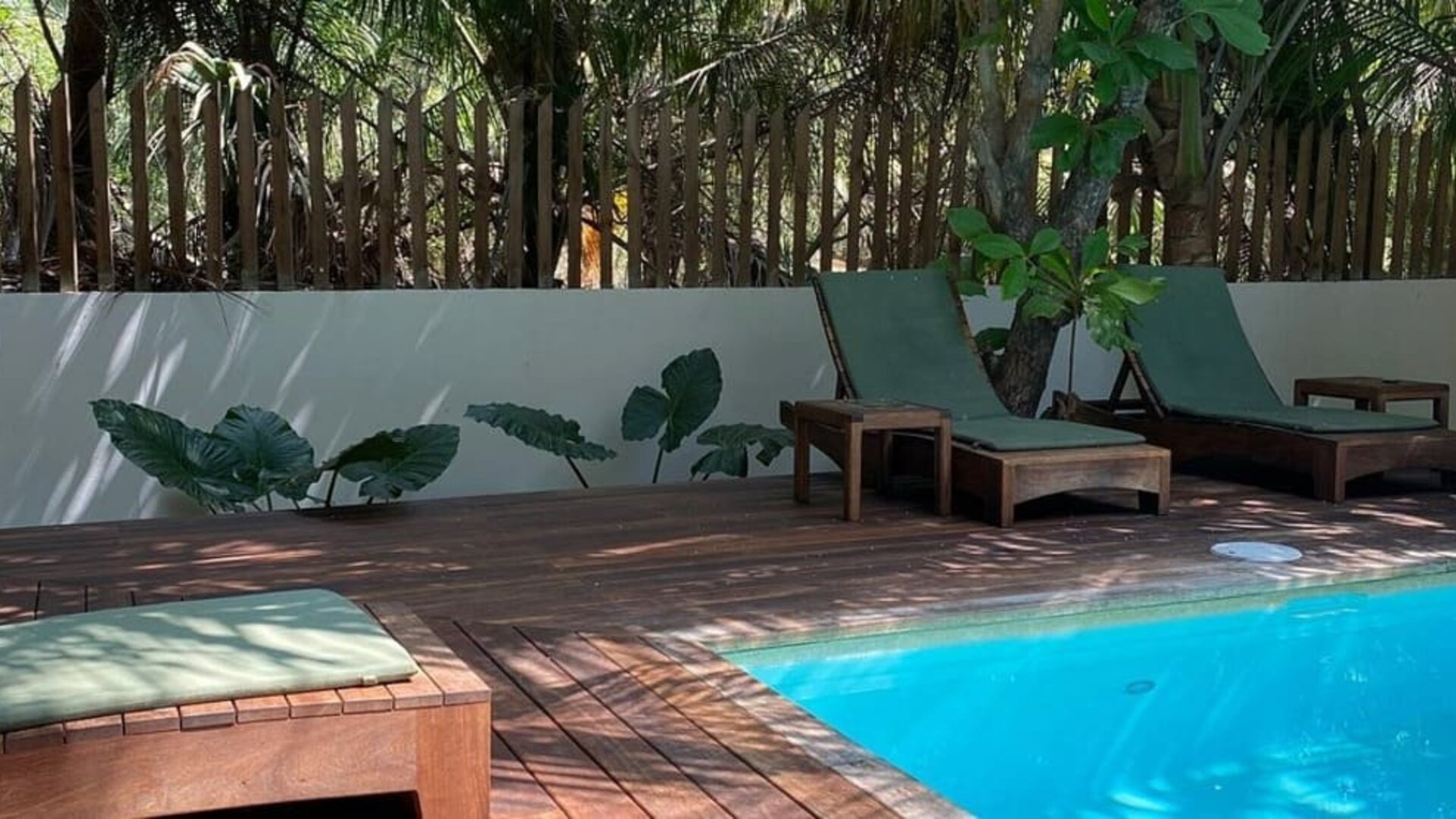 Shaded poolside area with wooden sun loungers and green cushions beside a bright turquoise swimming pool, surrounded by trees, tropical plants, and a wooden privacy fence.