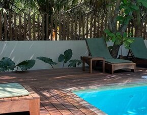 Shaded poolside area with wooden sun loungers and green cushions beside a bright turquoise swimming pool, surrounded by trees, tropical plants, and a wooden privacy fence.