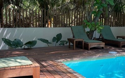 Shaded poolside area with wooden sun loungers and green cushions beside a bright turquoise swimming pool, surrounded by trees, tropical plants, and a wooden privacy fence.