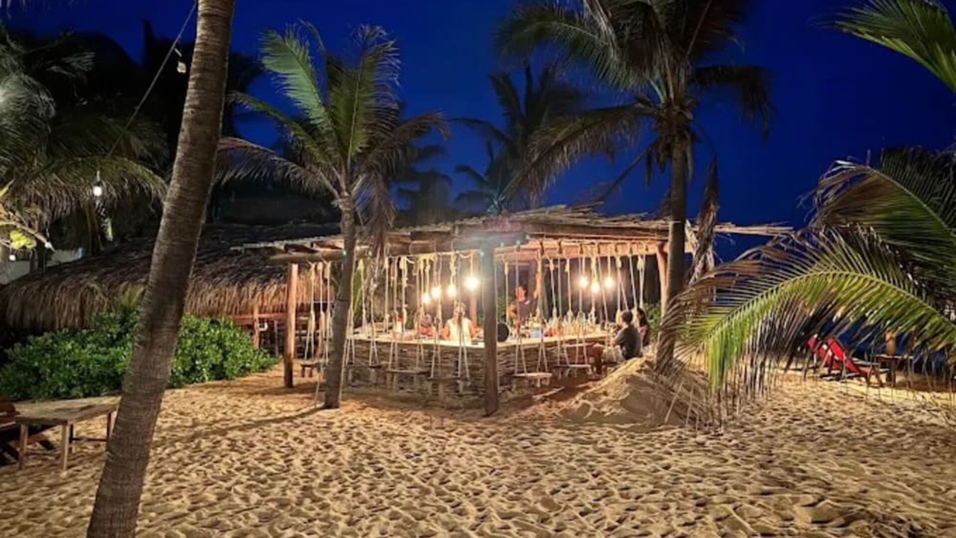 Illuminated open-air beach bar surrounded by tall palm trees and sandy grounds at night, creating a relaxed tropical atmosphere near the ocean.