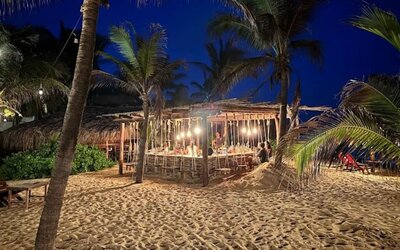 Illuminated open-air beach bar surrounded by tall palm trees and sandy grounds at night, creating a relaxed tropical atmosphere near the ocean.