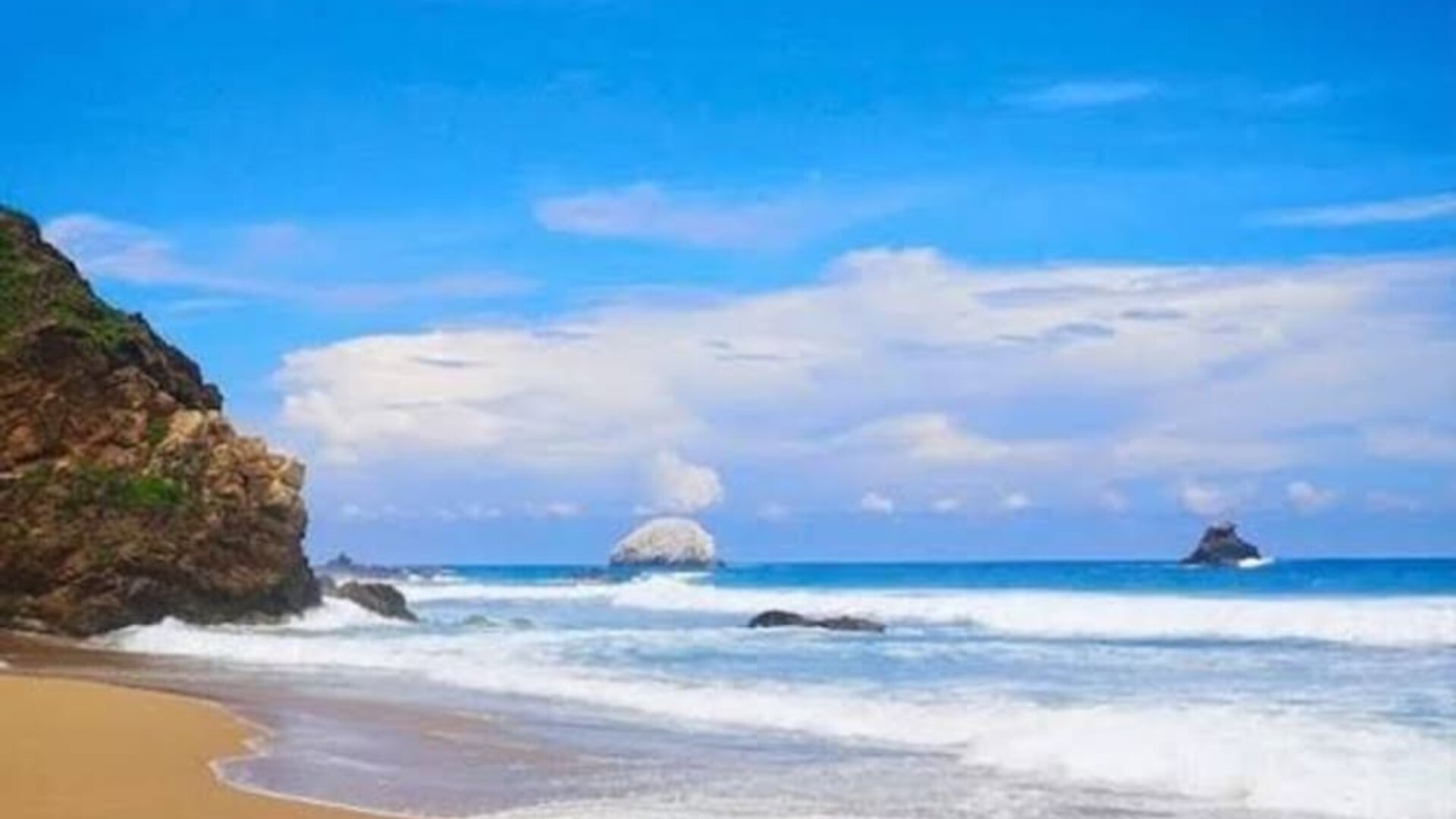 Scenic tropical coastline with golden sand, rolling ocean waves, rocky outcrops, and offshore rock formations beneath a bright blue sky with white clouds.