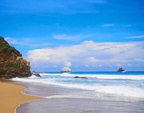 Scenic tropical coastline with golden sand, rolling ocean waves, rocky outcrops, and offshore rock formations beneath a bright blue sky with white clouds.