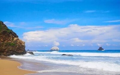 Scenic tropical coastline with golden sand, rolling ocean waves, rocky outcrops, and offshore rock formations beneath a bright blue sky with white clouds.