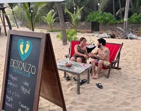 Descanso Beach Club sign beside two guests relaxing in red lounge chairs on the sand, surrounded by palm trees, tropical greenery, and casual beachfront seating.