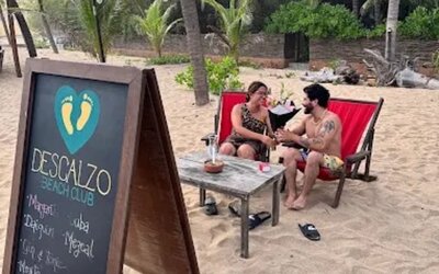 Descanso Beach Club sign beside two guests relaxing in red lounge chairs on the sand, surrounded by palm trees, tropical greenery, and casual beachfront seating.