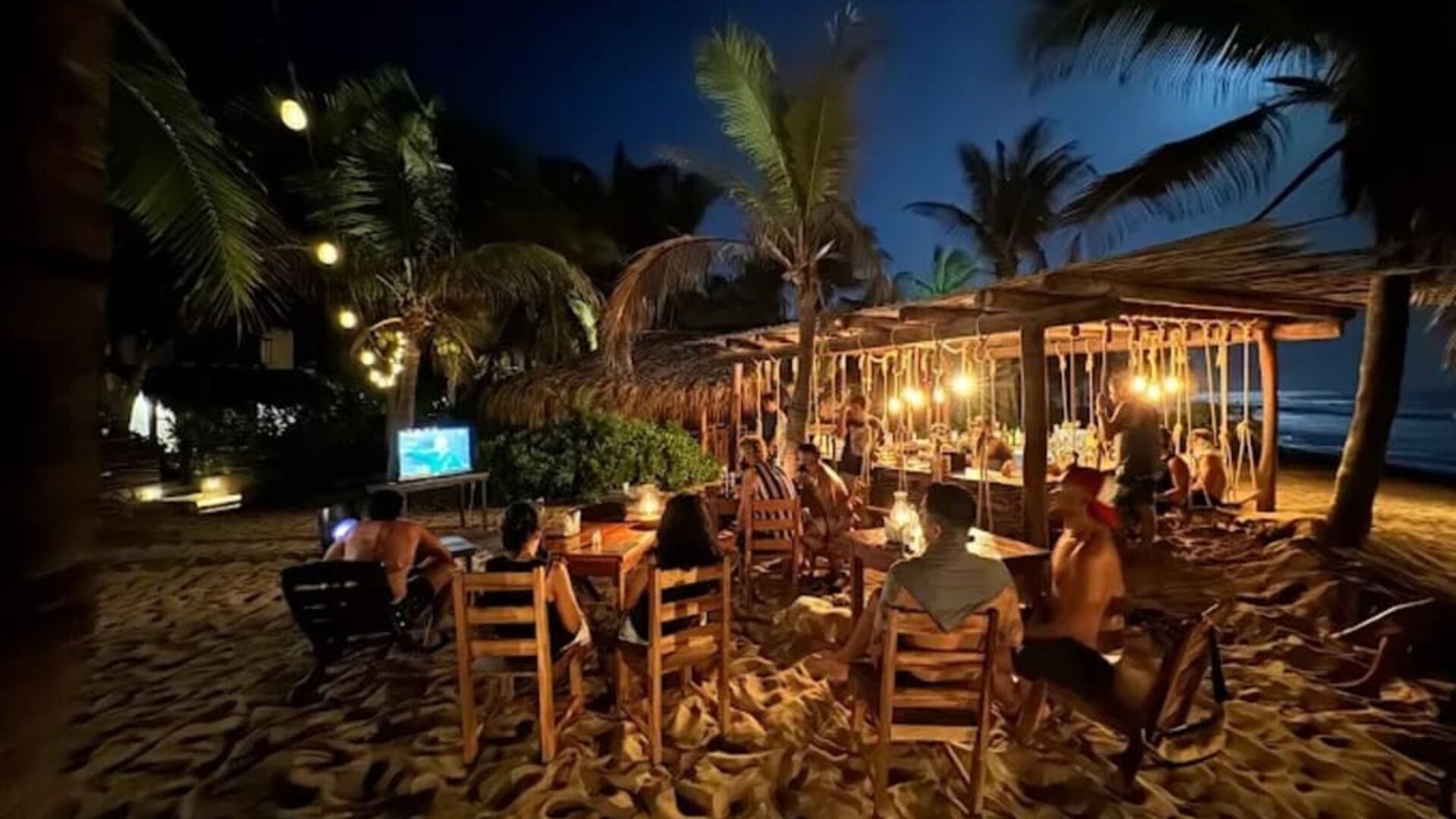 Guests sit at wooden tables on the sand beside a warmly lit beachfront bar, surrounded by palm trees with the dark ocean visible in the background.