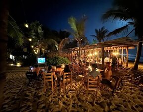 Guests sit at wooden tables on the sand beside a warmly lit beachfront bar, surrounded by palm trees with the dark ocean visible in the background.