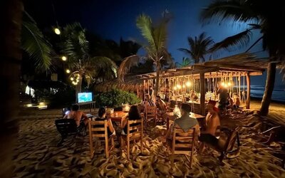 Guests sit at wooden tables on the sand beside a warmly lit beachfront bar, surrounded by palm trees with the dark ocean visible in the background.