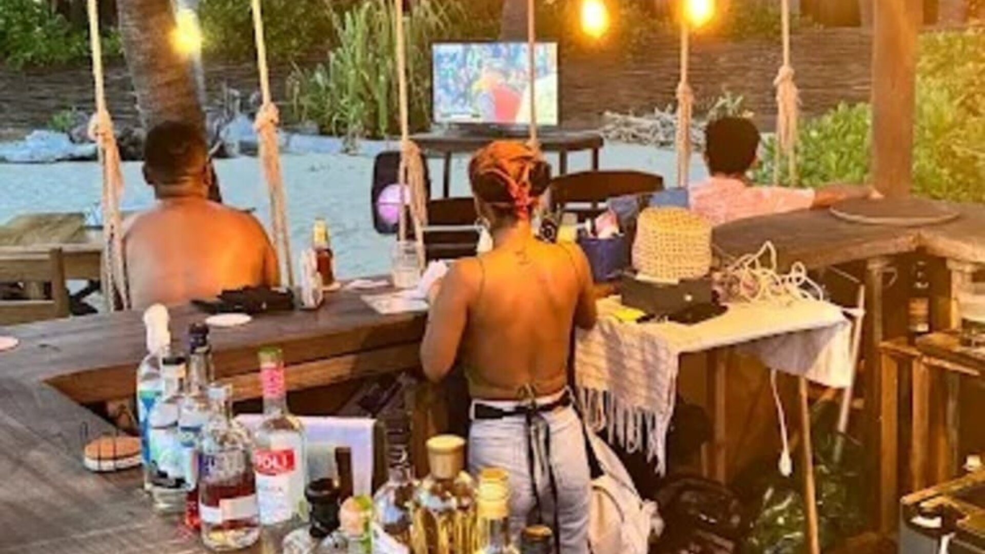 Rustic wooden beach bar with a large selection of liquor bottles, warm pendant lights, and staff behind the counter, with palm trees and an outdoor television beyond.