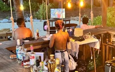 Rustic wooden beach bar with a large selection of liquor bottles, warm pendant lights, and staff behind the counter, with palm trees and an outdoor television beyond.