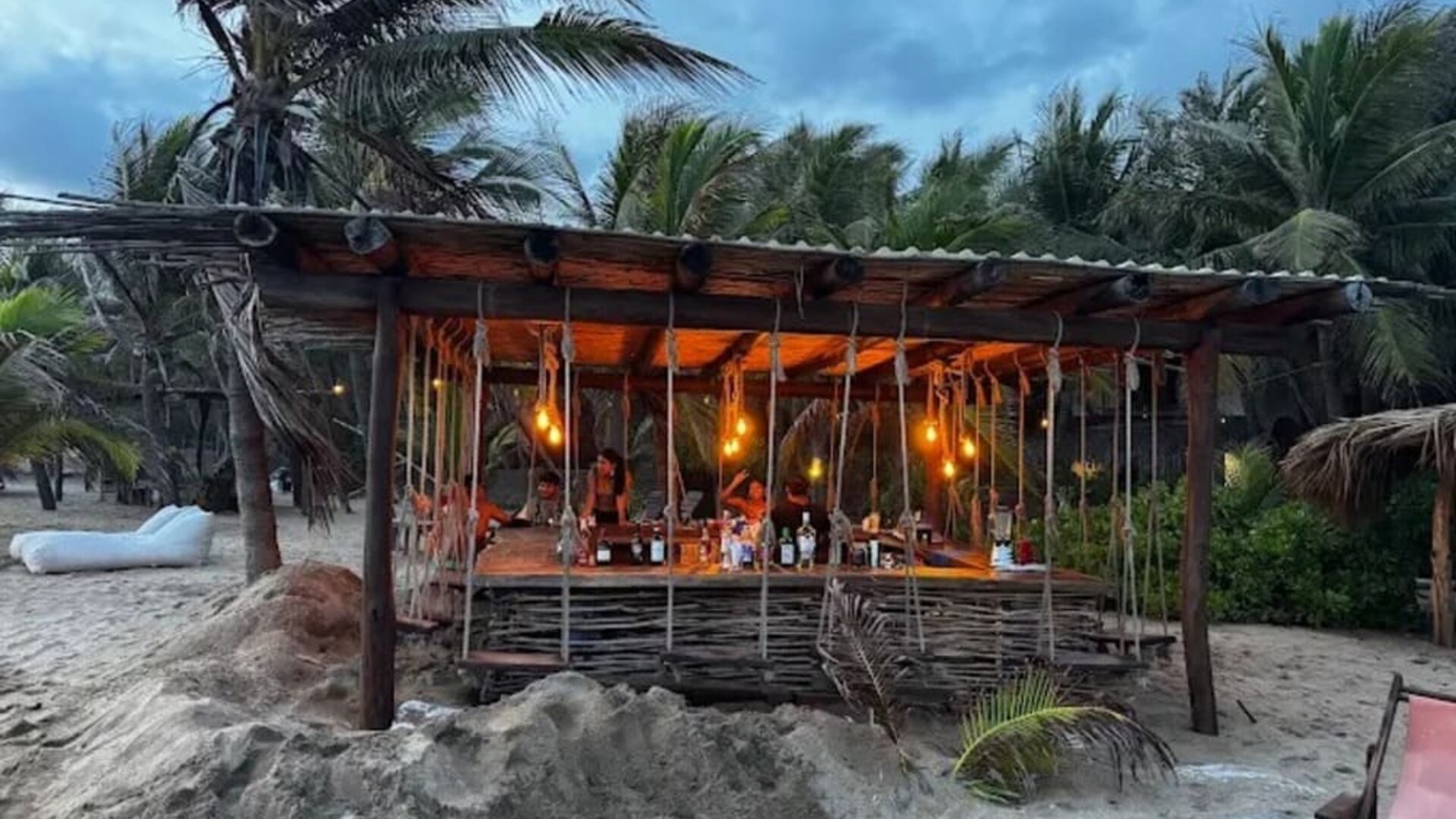 Rustic open-air beach bar built from wood and woven branches, illuminated by warm hanging lights and surrounded by sand, palm trees, and tropical vegetation.