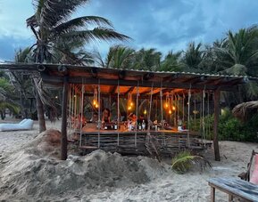Rustic open-air beach bar built from wood and woven branches, illuminated by warm hanging lights and surrounded by sand, palm trees, and tropical vegetation.