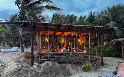 Rustic open-air beach bar built from wood and woven branches, illuminated by warm hanging lights and surrounded by sand, palm trees, and tropical vegetation.