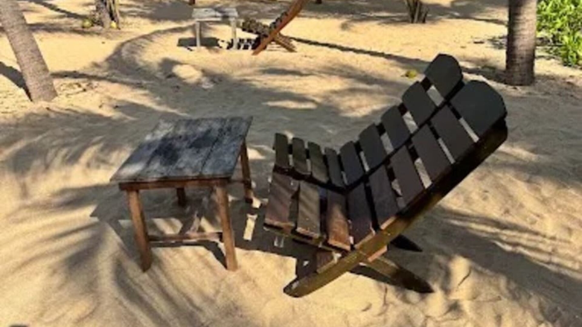 Wooden reclining beach chair and small side table positioned on the sand beneath palm trees, with shaded seating areas and the ocean visible nearby.