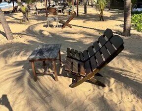 Wooden reclining beach chair and small side table positioned on the sand beneath palm trees, with shaded seating areas and the ocean visible nearby.