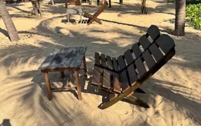 Wooden reclining beach chair and small side table positioned on the sand beneath palm trees, with shaded seating areas and the ocean visible nearby.