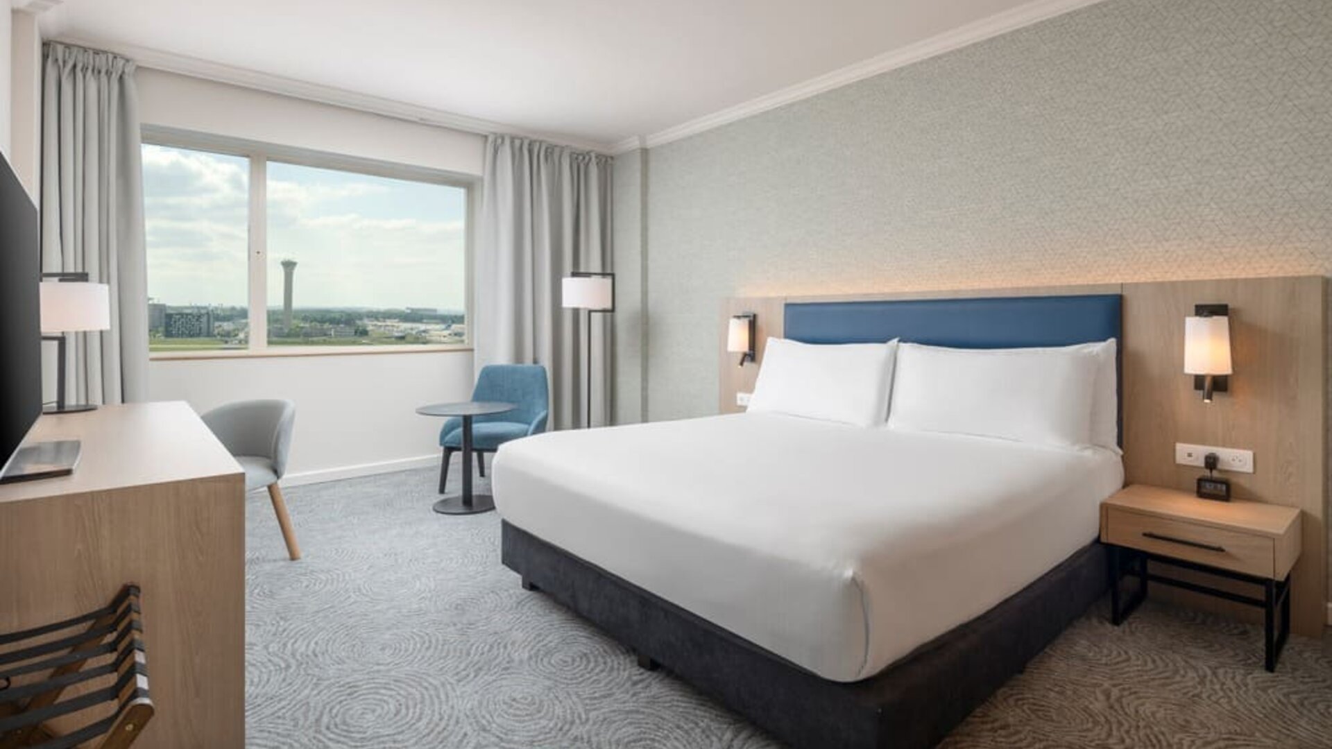 Modern king guest room with bed, large television, long work desk, armchairs, side table and floor-to-ceiling curtains.