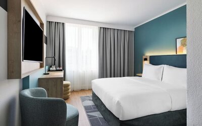 Bright contemporary hotel room with a king bed, white bedding, teal accent wall, large window with gray curtains, wall-mounted TV, desk, and upholstered armchair.