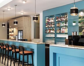 Contemporary hotel bar with turquoise counters, marble-look surfaces, high stools, pendant lighting, extensive bottle displays, glassware, and a coffee machine.