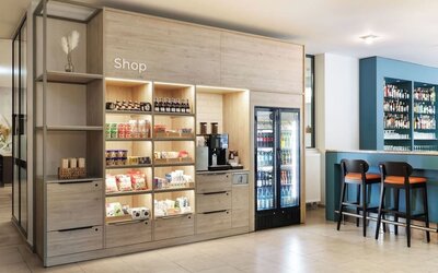 Hotel lobby shop with illuminated wooden shelves displaying snacks and drinks, a coffee machine, refrigerated beverages, and adjacent bar seating.