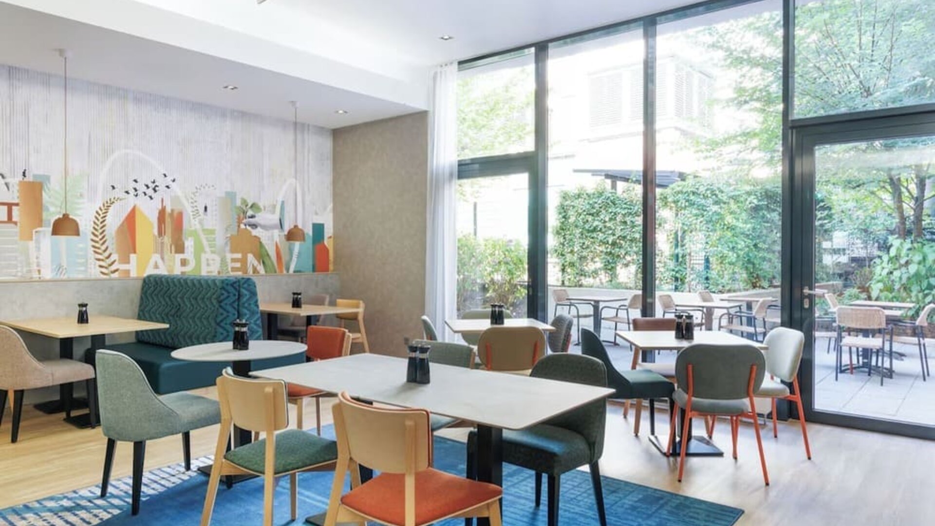 Contemporary hotel dining area with colorful upholstered chairs, wooden tables, teal booth seating, large floor-to-ceiling windows, and views of a leafy outdoor terrace.