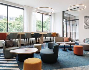 Spacious hotel lounge with teal, gray, orange, and yellow seating arranged around low tables, a blue patterned rug, large windows, and circular pendant lights.