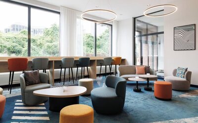 Spacious hotel lounge with teal, gray, orange, and yellow seating arranged around low tables, a blue patterned rug, large windows, and circular pendant lights.