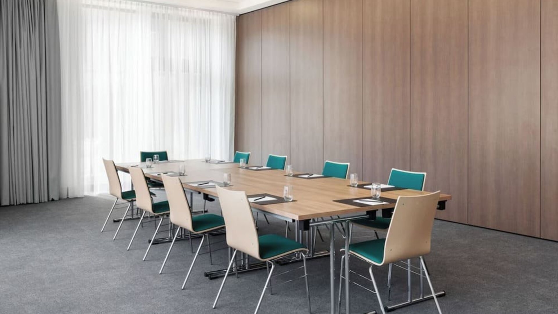 Bright hotel meeting room with rectangular tables arranged in a boardroom layout, teal and light wood chairs, water glasses and notepads, gray carpeting, and floor-to-ceiling curtains.