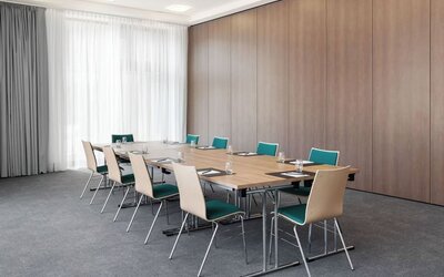 Bright hotel meeting room with rectangular tables arranged in a boardroom layout, teal and light wood chairs, water glasses and notepads, gray carpeting, and floor-to-ceiling curtains.