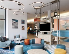 Contemporary hotel lobby with colorful lounge chairs and ottomans, round tables, blue patterned carpeting, circular ceiling lights, open shelving, and reception desk in the background.