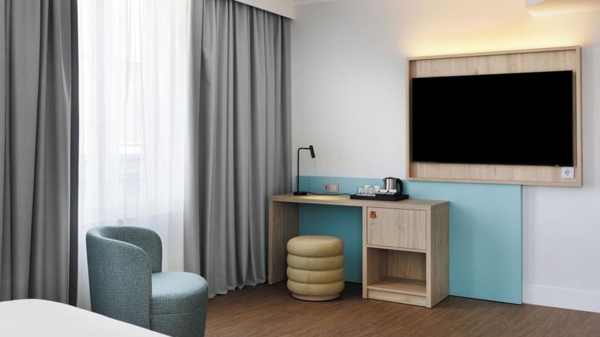 Hotel guest room workspace with wall-mounted TV, light wood desk, black reading lamp, electric kettle with cups, round upholstered stool, armchair, and floor-to-ceiling curtains.