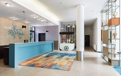 Modern hotel reception area with a turquoise front desk, colorful abstract rug, decorative open shelving, white column, hydration station, and Spark branding on a wood-paneled wall.