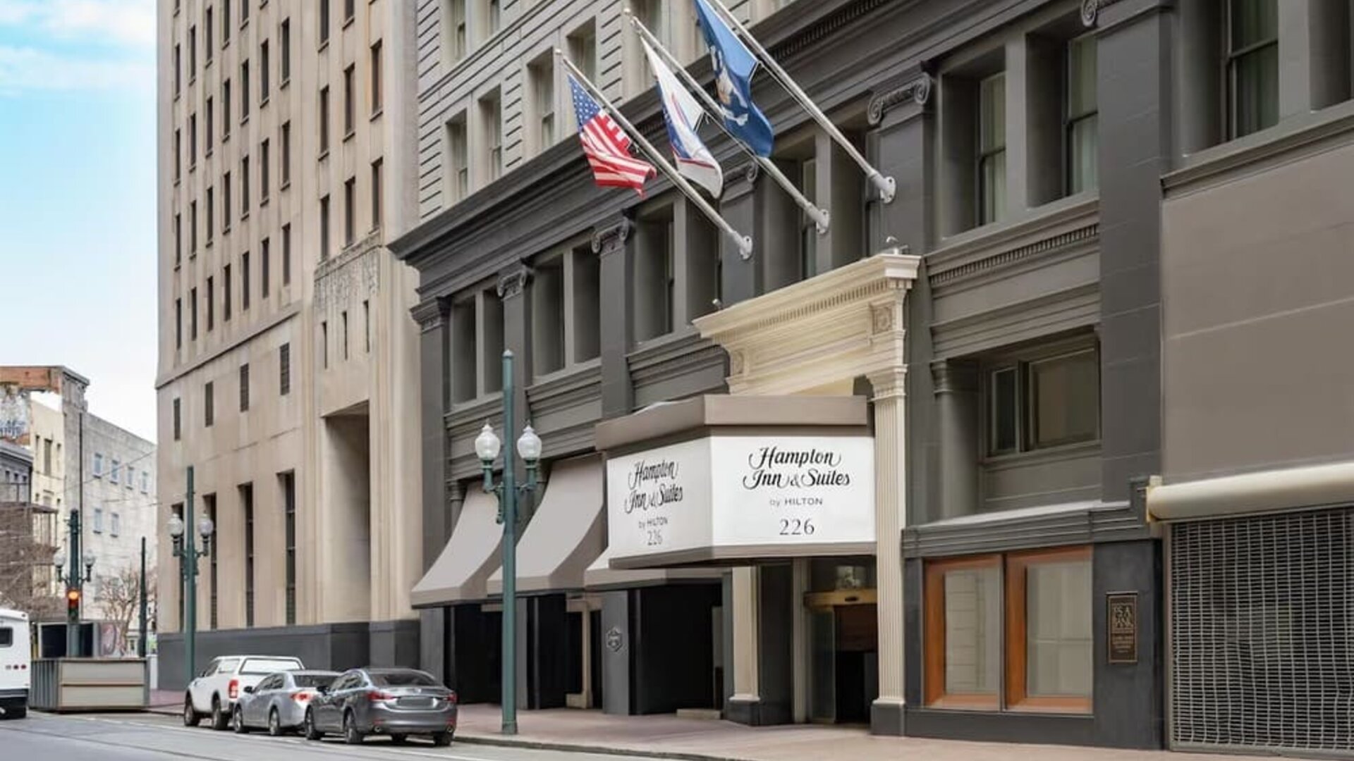 Street-level view of a multi-story urban hotel with a dark gray façade, columned entrance canopy, American and other flags above the doorway, and parked cars along the curb.