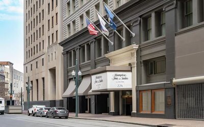 Street-level view of a multi-story urban hotel with a dark gray façade, columned entrance canopy, American and other flags above the doorway, and parked cars along the curb.