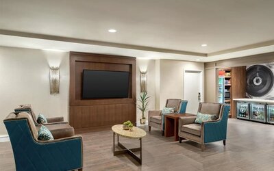 Comfortable hotel lounge with teal and gray upholstered armchairs, patterned cushions, oval coffee table, wall-mounted flat-screen TV, decorative lighting, and access to the adjacent convenience shop.