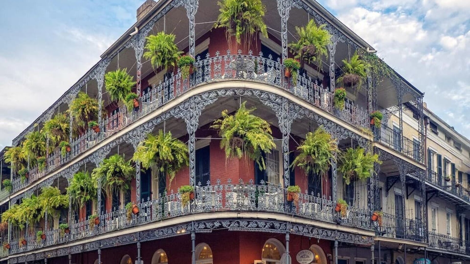 Historic New Orleans corner building with red exterior walls, ornate wrought-iron balconies on multiple levels, abundant hanging ferns, ground-floor storefronts, bicycles, and pedestrians along the street.