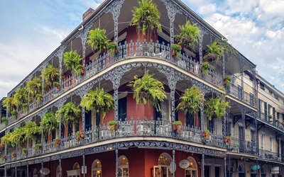 Historic New Orleans corner building with red exterior walls, ornate wrought-iron balconies on multiple levels, abundant hanging ferns, ground-floor storefronts, bicycles, and pedestrians along the street.