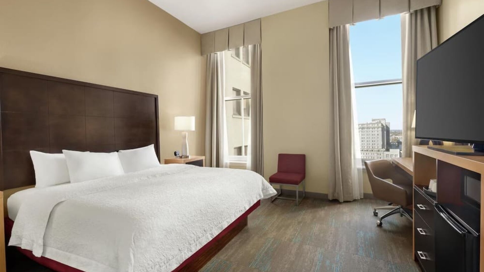 Bright hotel guest room with a king bed, white linens, dark wood headboard, tall windows with neutral curtains, bedside lamp, desk and chair, flat-screen TV, and city views.